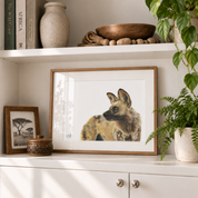 A framed art print of a spotted hyena with a light brown wood frame and white mat, displayed on a white shelf next to a small framed print, a decorative box, and a potted plant.