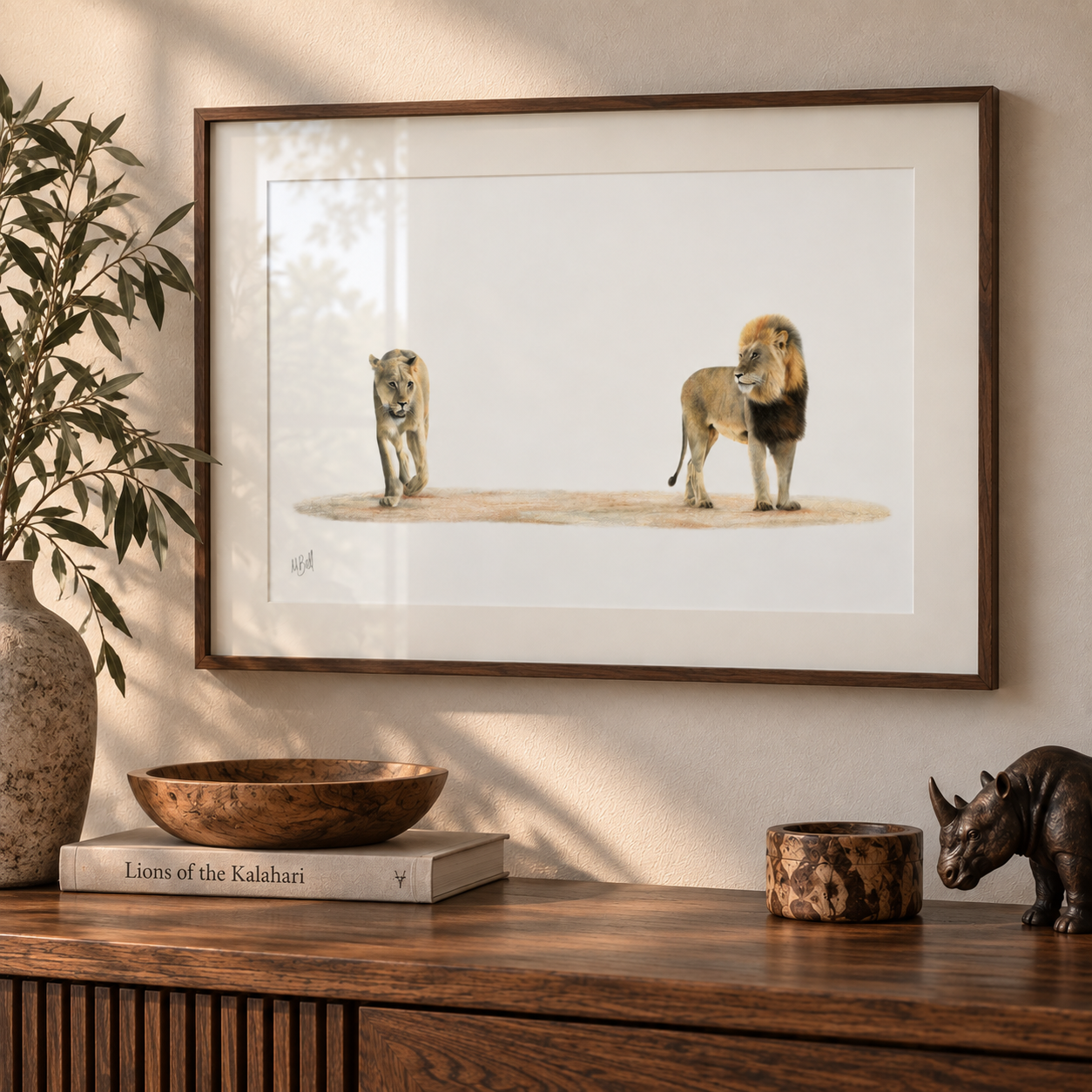 A framed art print depicting a lioness and a male lion standing on a dry patch of ground, displayed on a wooden console table in a styled room setting with a plant, books, and decor.
