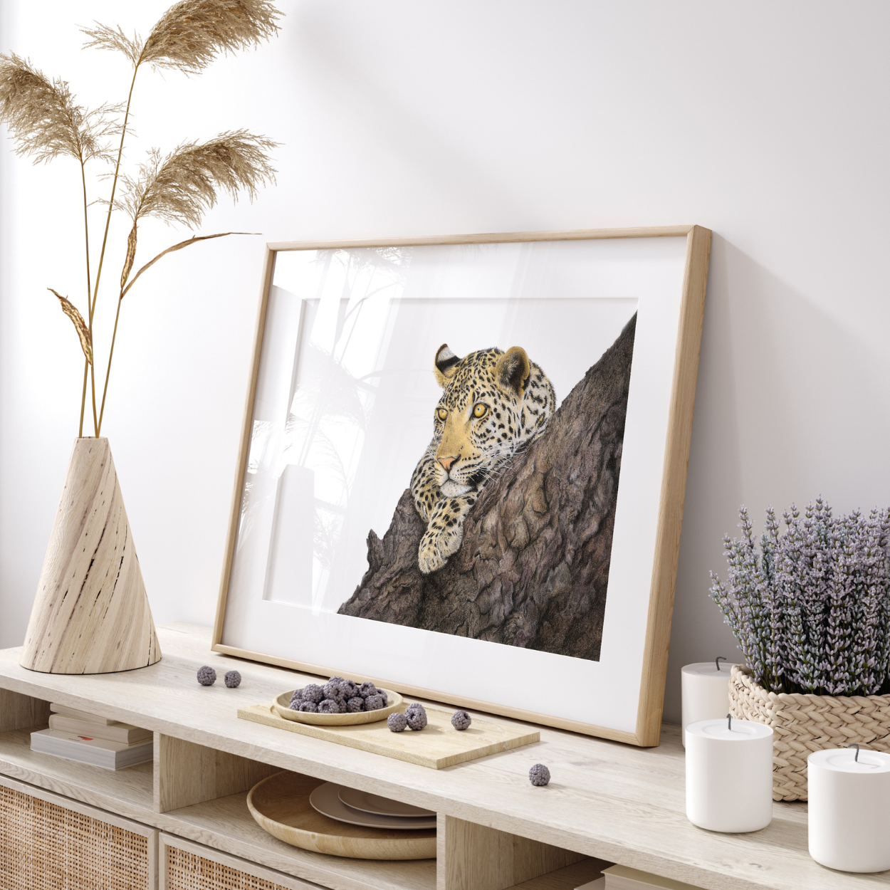 A framed art print of a leopard resting on a tree branch, styled on a light wood console table with dried grass in a vase, candles, and small plants.