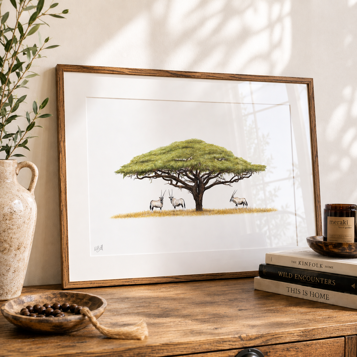 A framed art print featuring a detailed illustration of an acacia tree with three gemsbok, styled on a wooden table next to a ceramic vase and books.