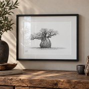 A framed black and white pencil sketch of a baobab tree, displayed on a rustic wooden table in a warm, styled interior setting with a potted olive and small ceramic vessels.