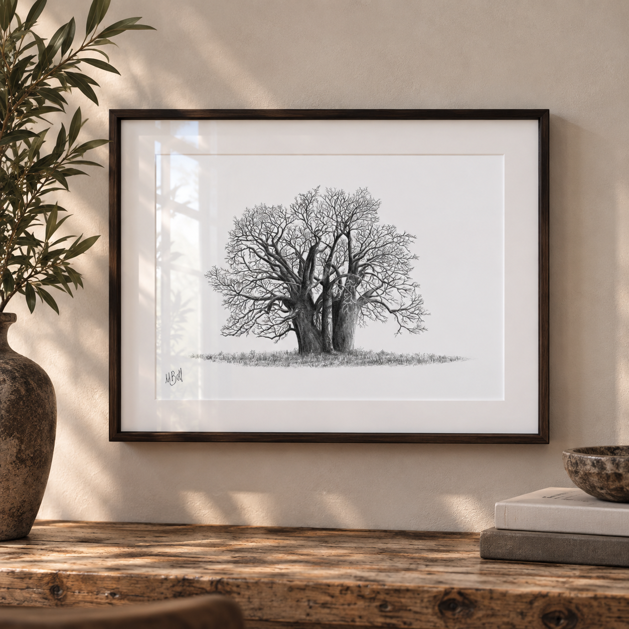 A framed black and white botanical art print of two ancient trees, displayed on a rustic wooden shelf next to a potted plant and books against a warm beige wall.