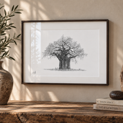 A framed black and white sketch of a baobab tree on a white mat, displayed on a rustic wooden table against a beige wall with decorative plants and books.