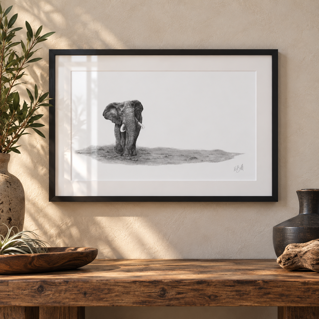 A framed black and white pencil drawing of an elephant on a white mat, hung on a textured beige wall above a rustic wooden table with decorative vases and a bowl.