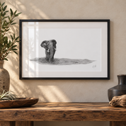 A framed black and white pencil drawing of an elephant on a white mat, hung on a textured beige wall above a rustic wooden table with decorative vases and a bowl.