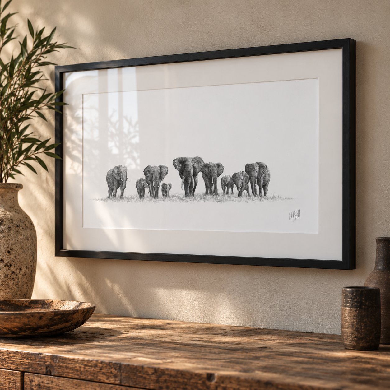 A framed black and white pencil sketch of a herd of elephants, displayed on a beige wall above a rustic wooden table with decorative ceramic vases.