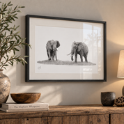 A framed black and white elephant art print hangs on a beige wall above a rustic wooden console table styled with a vase, books, and a lamp.