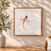A framed art print featuring an African Paradise Flycatcher with a long tail perched on a branch with small red berries, displayed on a wall in a styled interior setting with a vase of leaves and a woven chair partially visible.