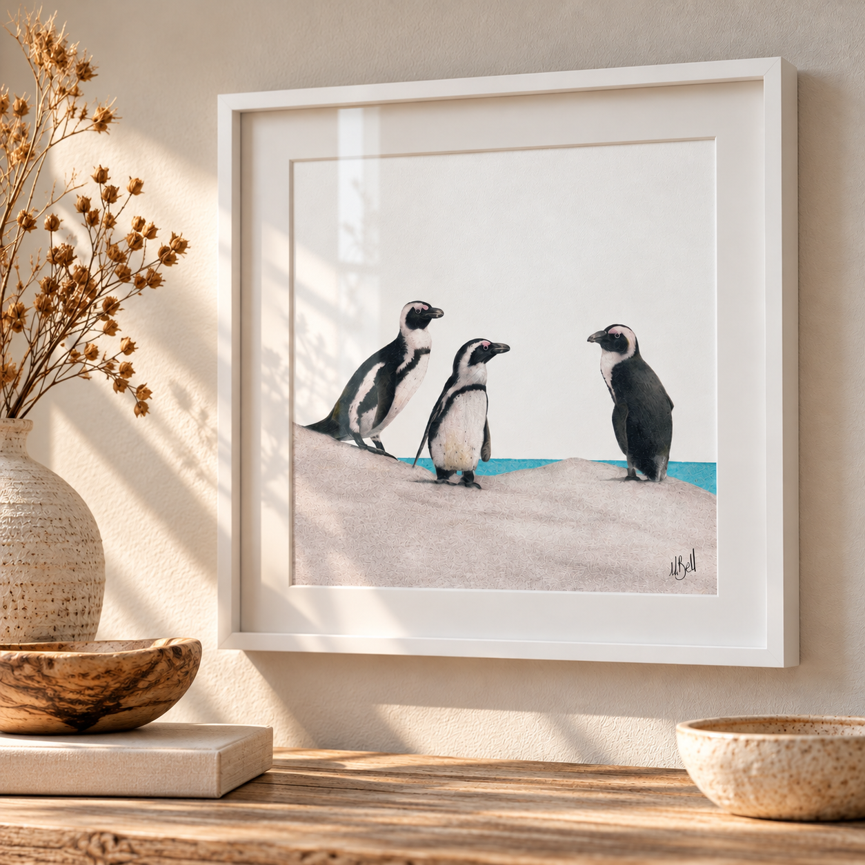 A framed art print featuring three african penguins standing on a sandy beach with blue water, displayed on a wooden table next to a vase with dried flowers.