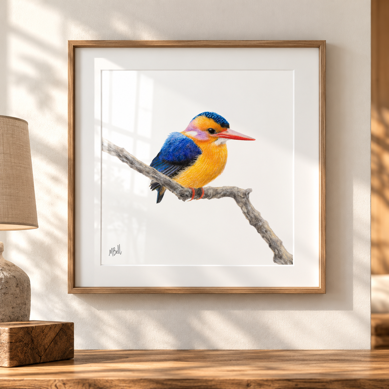 A framed art print of an African Pygmy Kingfisher perched on a branch, displayed on a wooden table against a white wall with soft window shadows. A lamp is visible on the left.