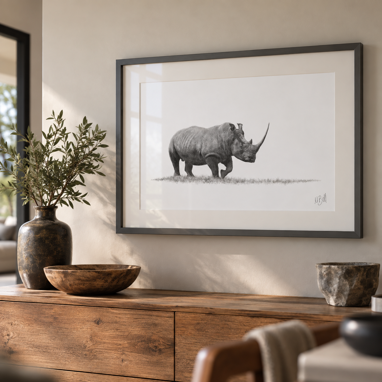 A framed black and white sketch print of a rhinoceros hanging on a beige wall above a wooden console table with decorative bowls and a vase.