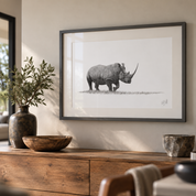A framed black and white sketch print of a rhinoceros hanging on a beige wall above a wooden console table with decorative bowls and a vase.