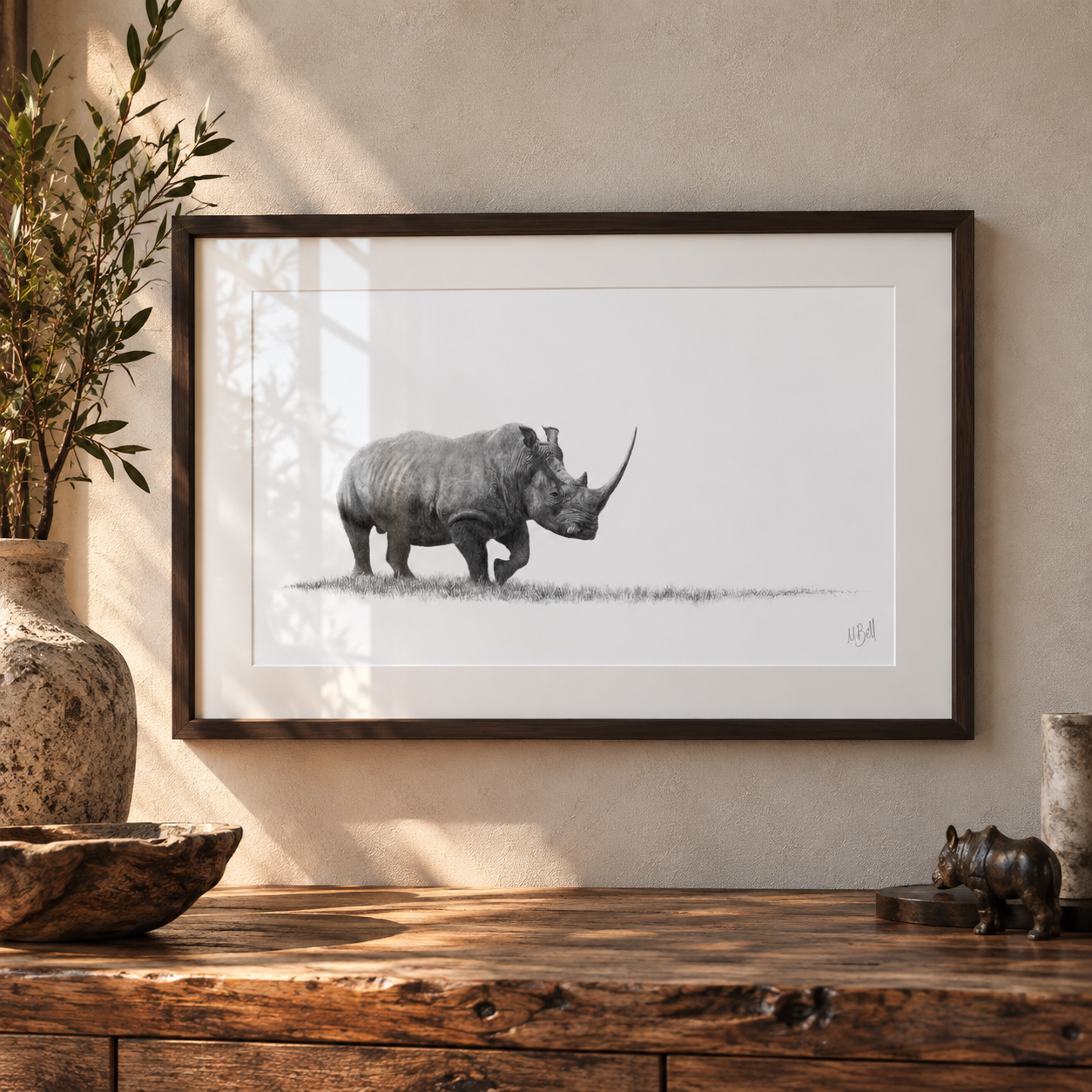 A framed black and white art print of a rhinoceros walking, displayed on a textured beige wall above a rustic wooden table with decorative props.