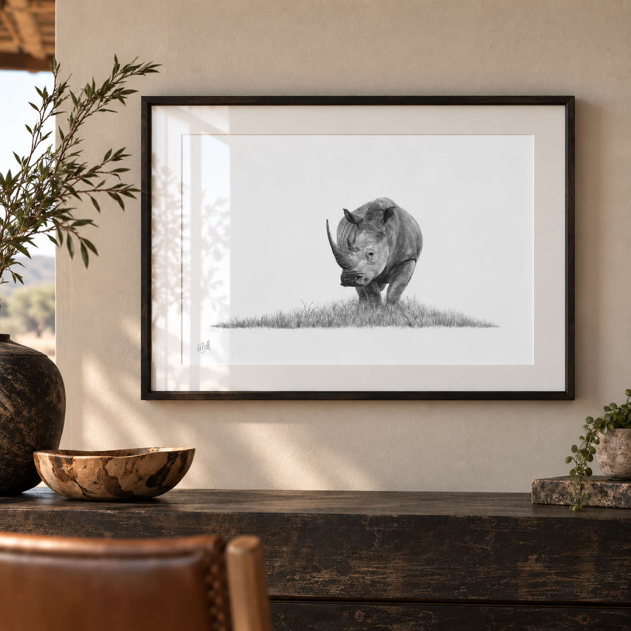 A framed black and white art print of a rhinoceros on a white background, hanging on a beige wall above a dark wooden console table with decorative plants and a bowl.