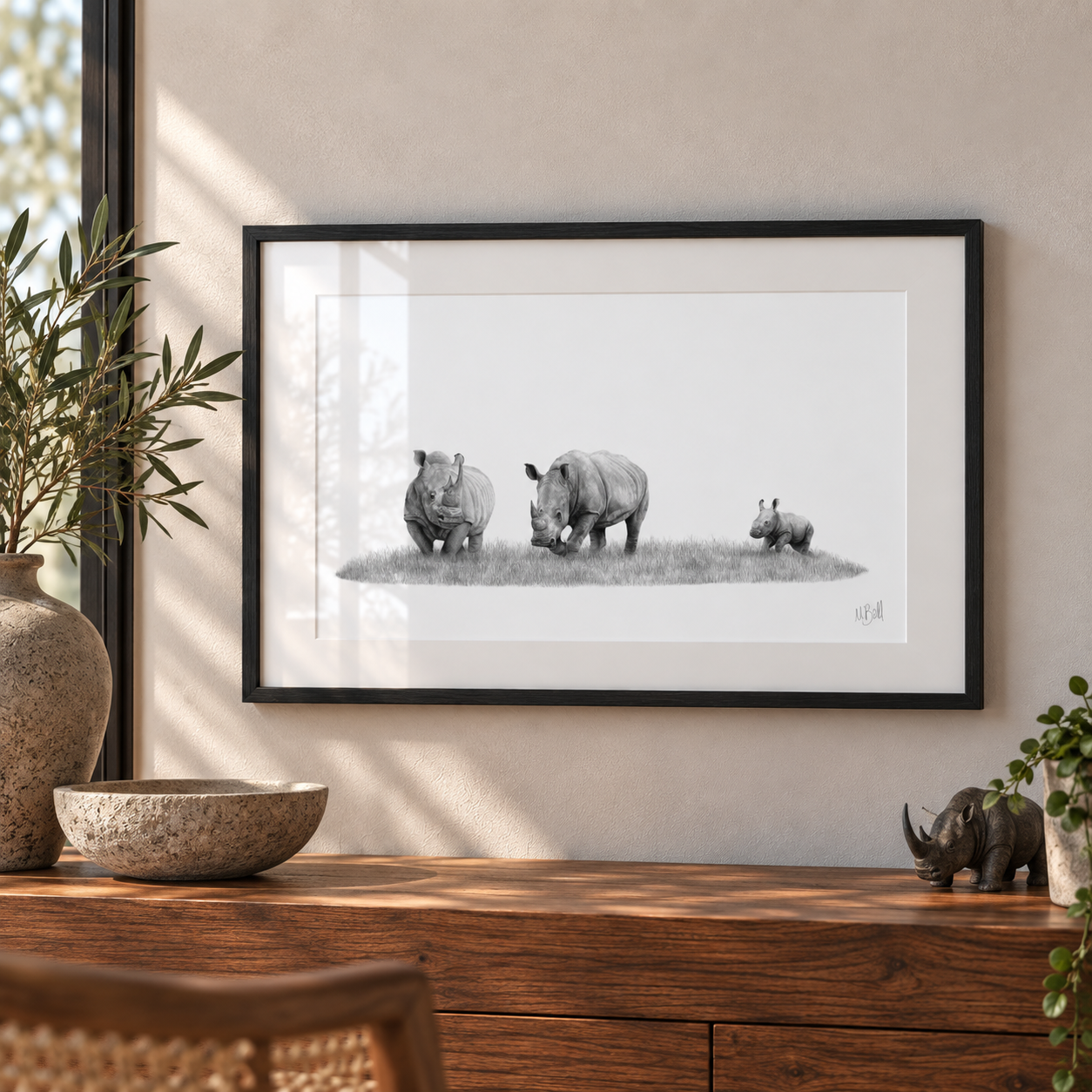 A framed black and white art print depicting a rhinoceros family, displayed on a beige wall above a wooden console table with decorative plants and a small rhino figurine.
