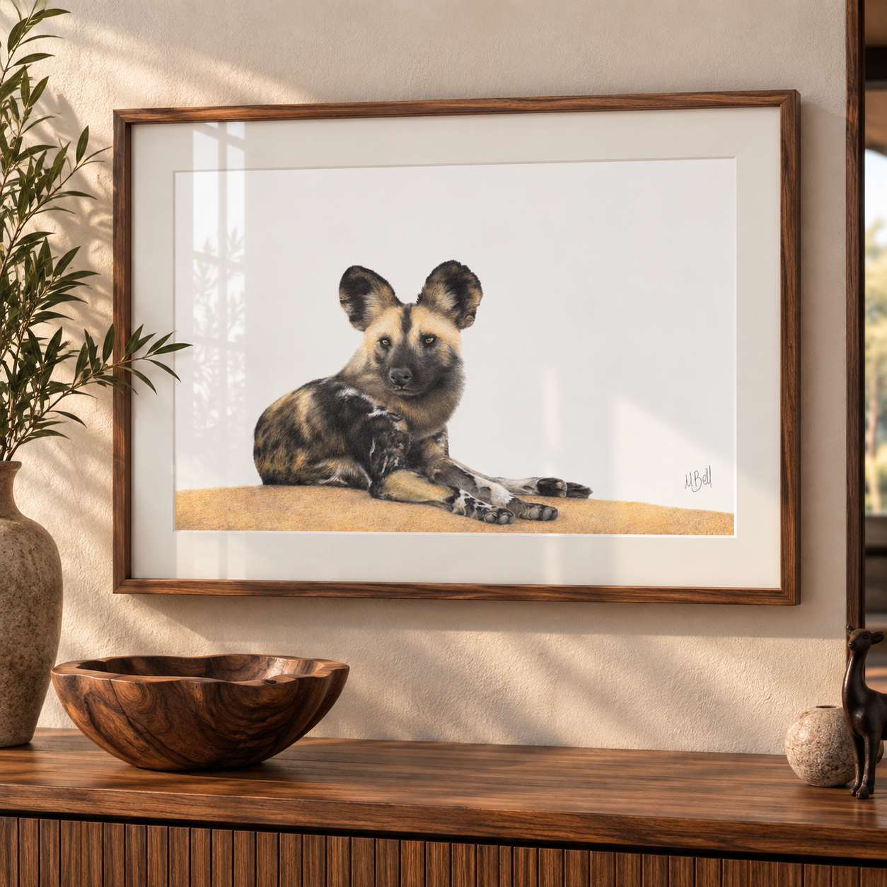 Framed wall art featuring a painted wild dog resting on a tan surface, displayed in a warm wood frame on a beige wall above a wooden console table with a bowl and plant.