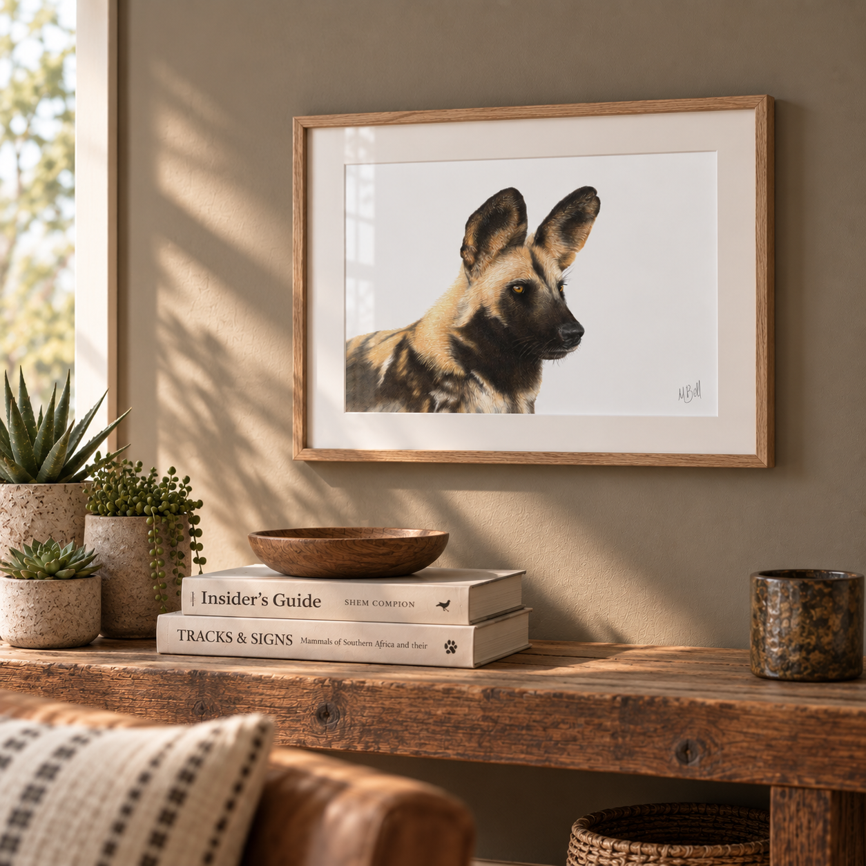 A framed art print of an African wild dog hanging on a warm beige wall above a rustic wooden console table styled with books, plants, and a bowl.
