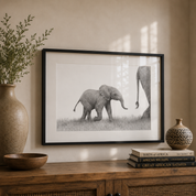 A framed black and white pencil sketch by South African Wildlife artist Matthew Bell of an elephant calf standing in grass, displayed on a wooden console table in a styled room setting with a vase, bowl, and decorative books.