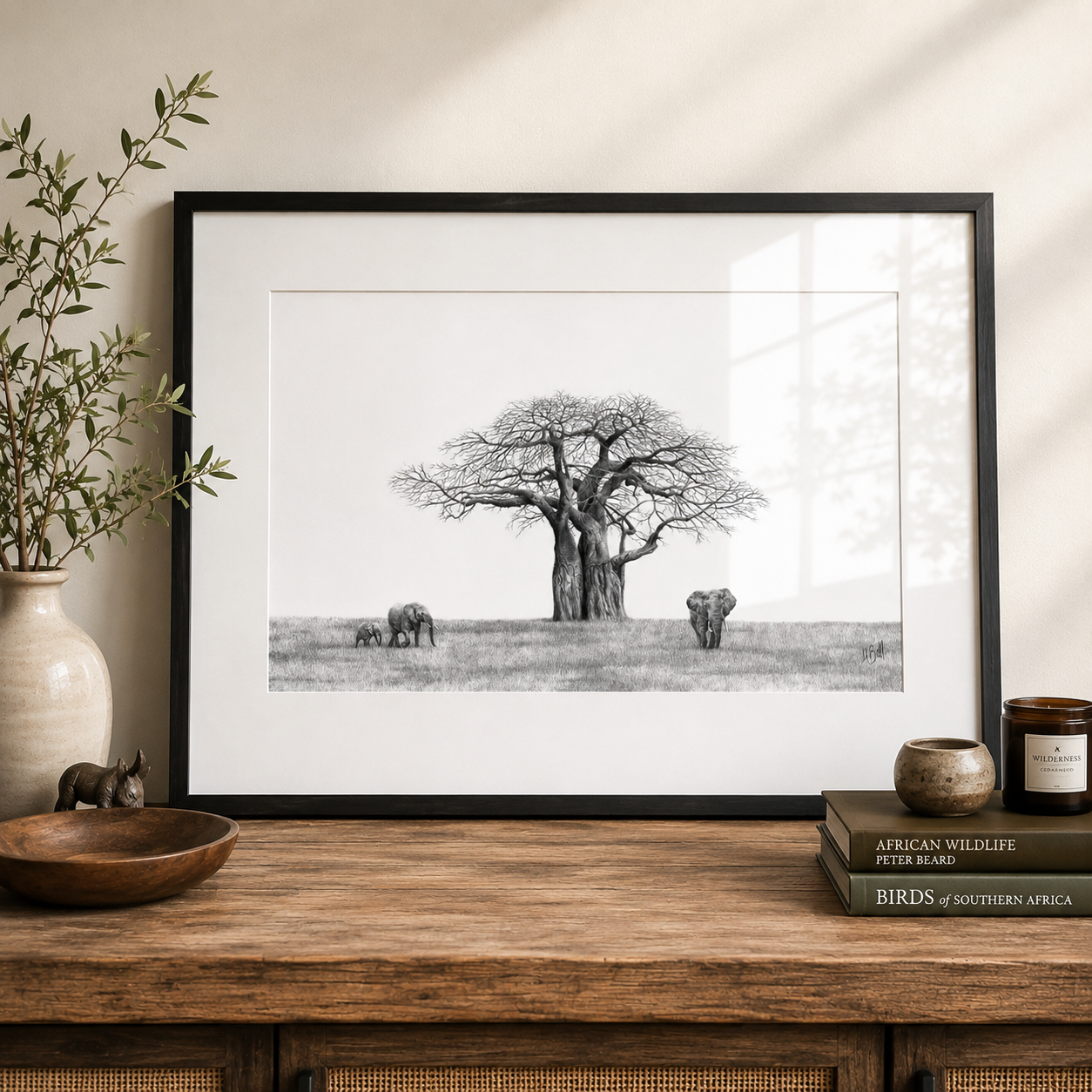 A framed black and white wildlife art print by artist Matthew Bell depicting a baobab tree and elephants, styled on a wooden console table with a vase, bowl, and books.