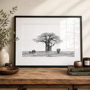 A framed black and white wildlife art print by artist Matthew Bell depicting a baobab tree and elephants, styled on a wooden console table with a vase, bowl, and books.