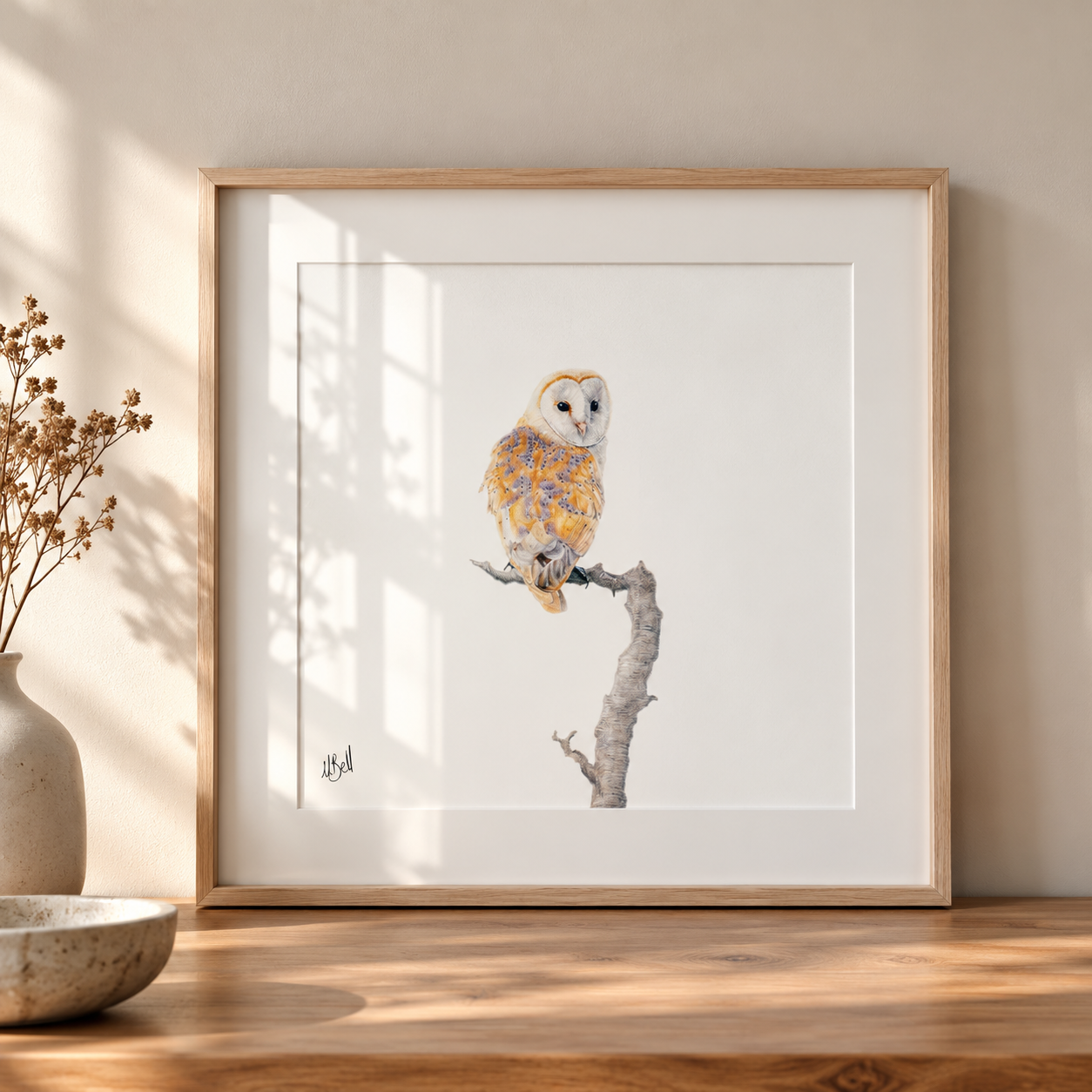 A framed pencil art print of a barn owl perched on a branch, displayed on a wooden table against a beige wall with soft window shadows.
