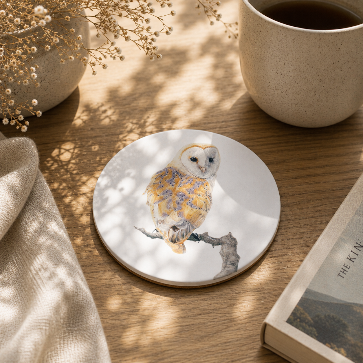 A round white coaster featuring a detailed illustration of a barn owl perched on a branch, decorated with yellow and blue floral patterns on its feathers. The coaster is styled on a wooden table next to a ceramic mug, a book, and dried flowers.