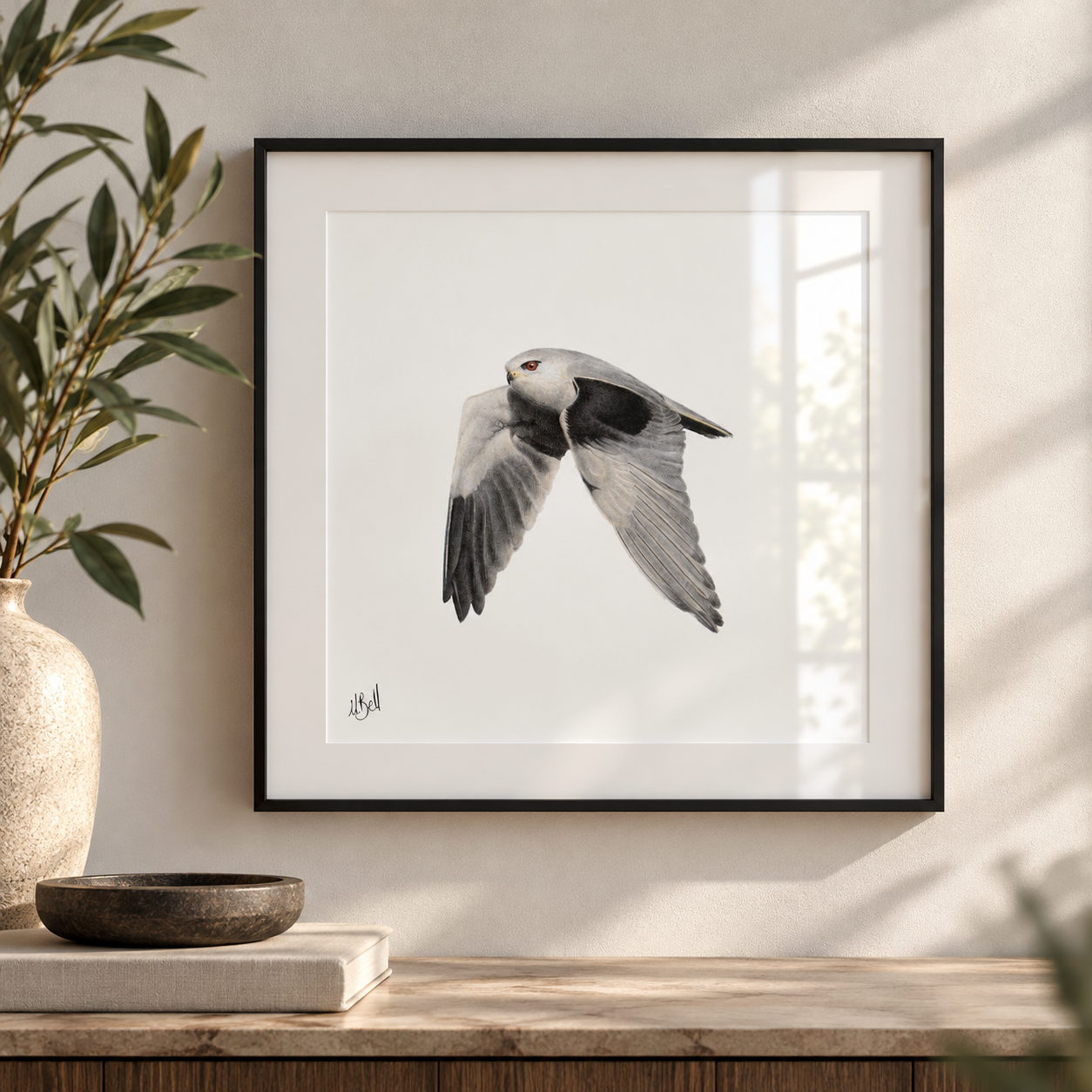 A framed art print of a Black Winged Kite in flight, displayed on a light beige wall above a wooden console table with a vase and bowl.