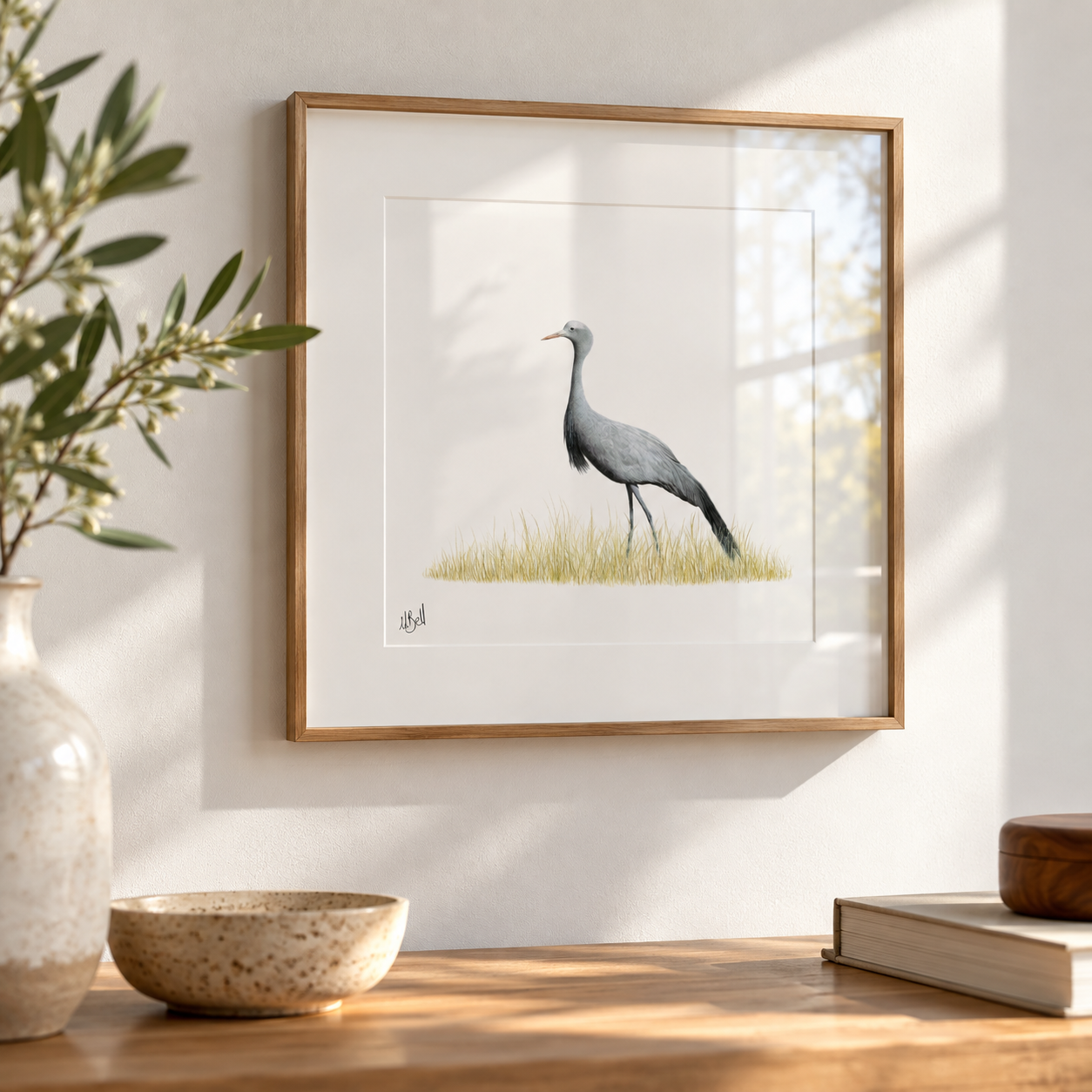 Framed wall art featuring a Blue Crane standing in a patch of yellow grass, displayed on a light wall above a wooden surface with a vase, bowl, and books nearby.