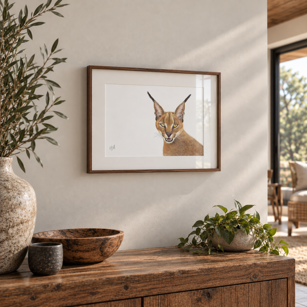 A framed art print of a caracal cat's face, displayed on a warm beige wall above a rustic wooden console table decorated with a ceramic vase, bowl, and potted plant.