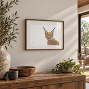 A framed art print of a caracal cat's face, displayed on a warm beige wall above a rustic wooden console table decorated with a ceramic vase, bowl, and potted plant.