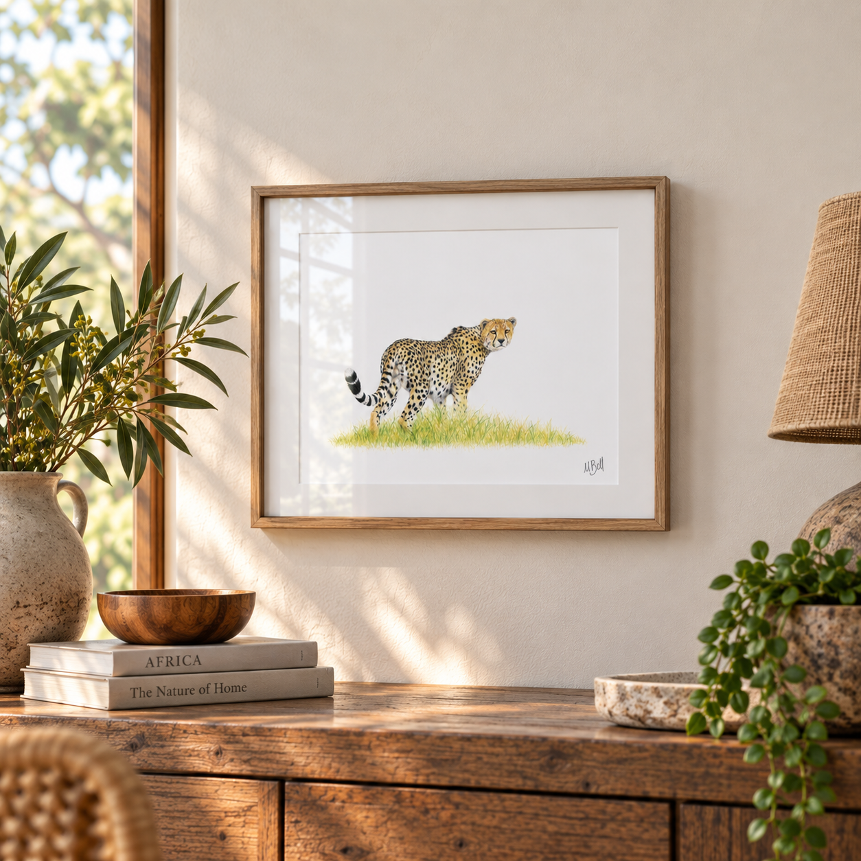 Framed wall art featuring a watercolor-style cheetah standing on grass, displayed in a warm, sunlit room above a wooden console table with books, plants, and a lamp.