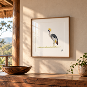 A framed art print of a crowned crane hanging on a beige plaster wall in a rustic room setting with a wooden bowl and plant.