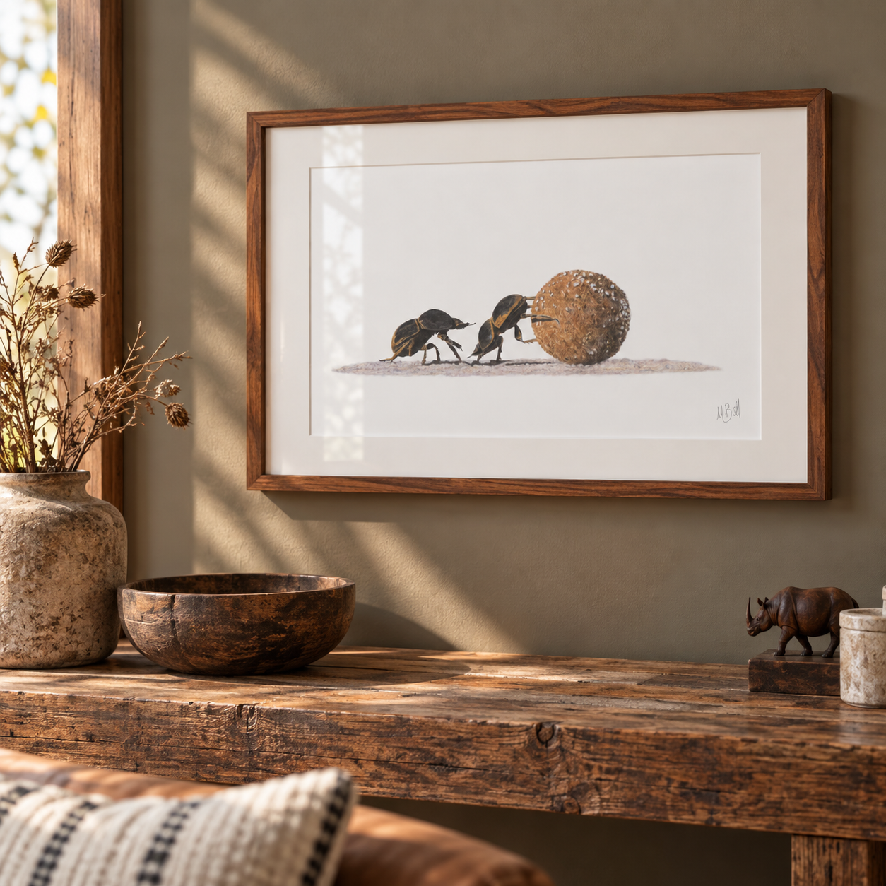 A framed art print featuring an illustration of two small dung beetles pushing a large round ball of dung, displayed on a rustic wooden console table in a warm, styled interior setting.