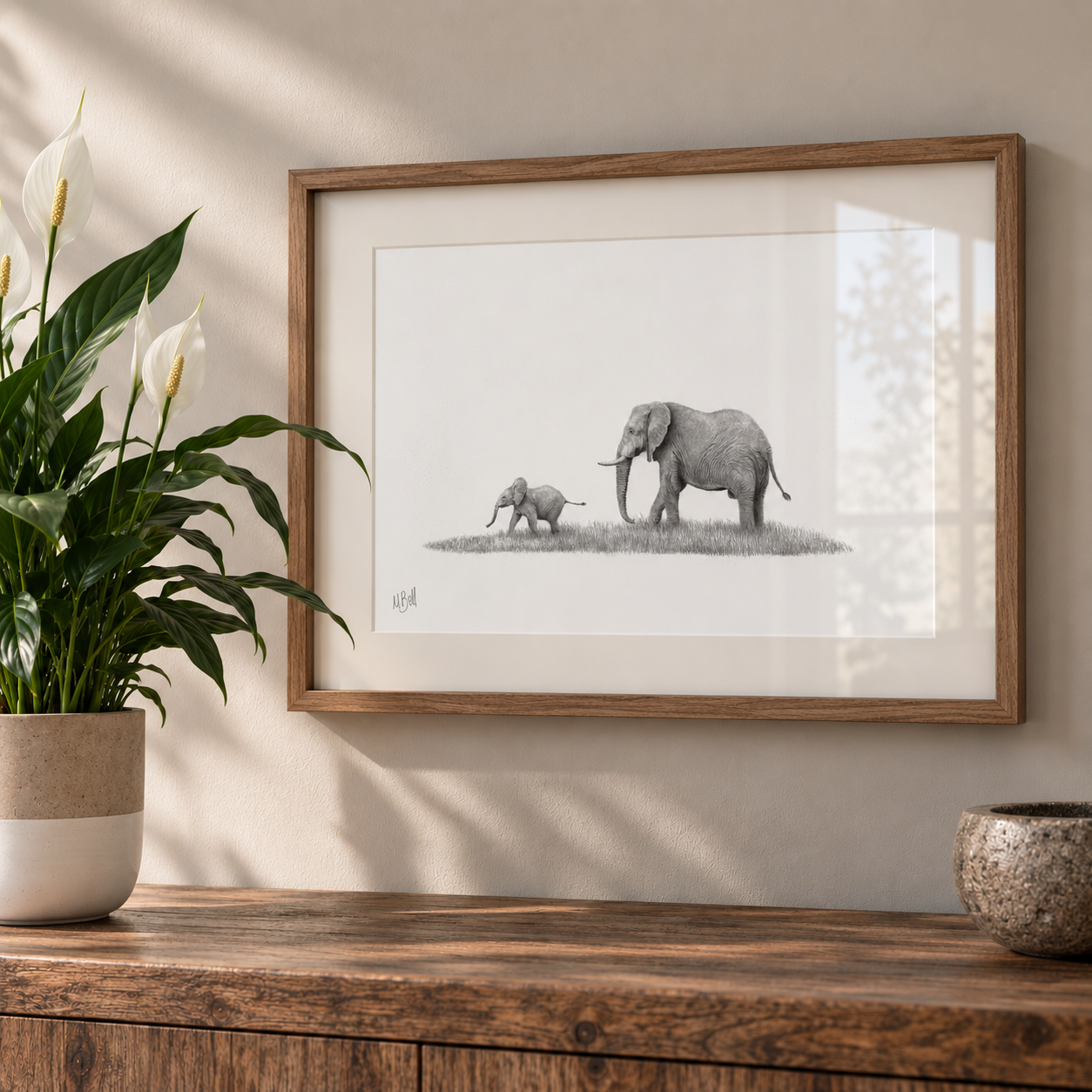 A framed black and white art print depicting an adult elephant and a baby elephant standing on grass, hung on a beige wall above a wooden console table with a potted plant and a bowl.