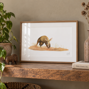 A framed art print featuring a painted aardvark on a small patch of grass, displayed on a rustic wooden shelf in a styled home setting with a plant, small elephant figurine, and book.