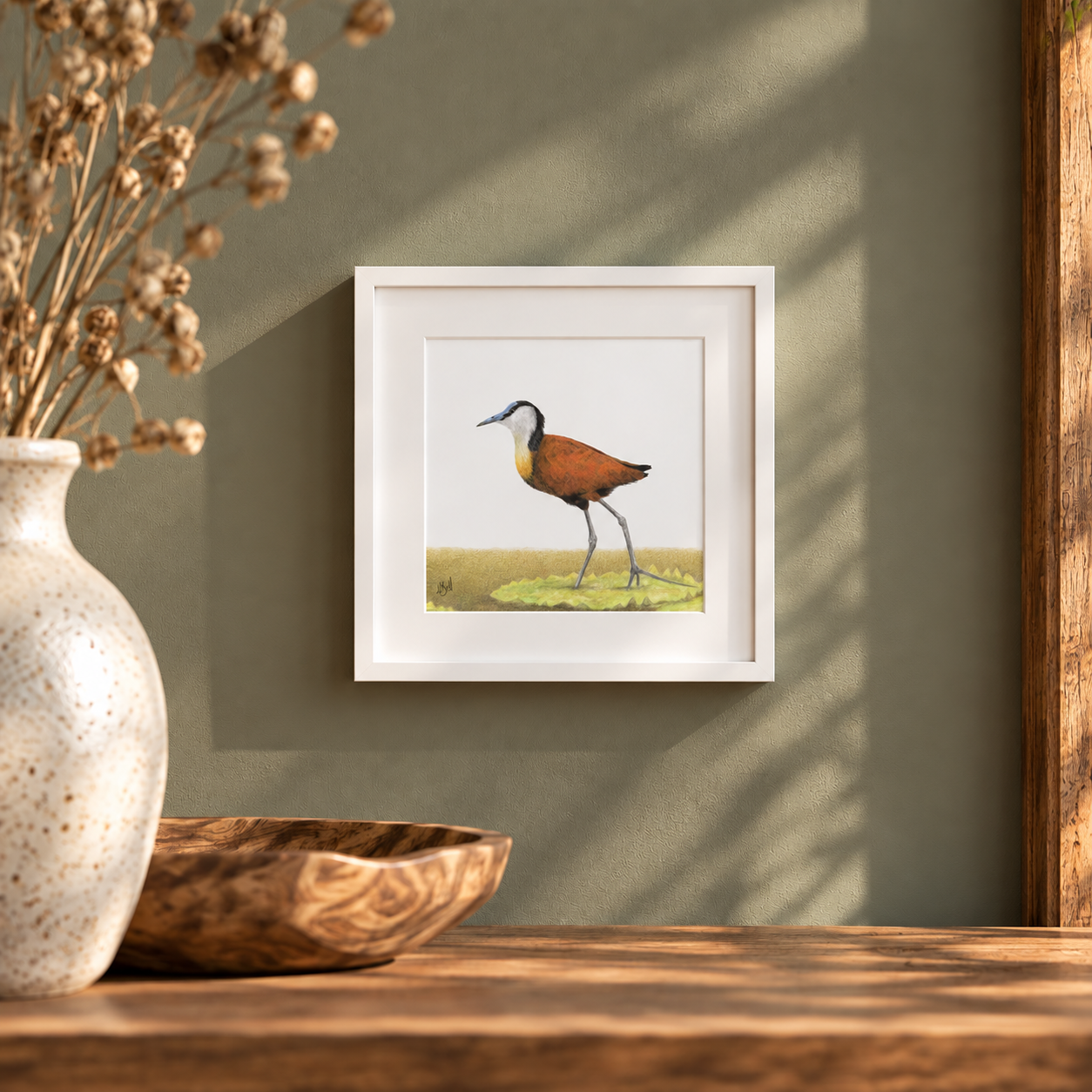 A framed art print of a bird standing on a lily pad, displayed on a dark green wall above a wooden table with a ceramic vase and a wooden bowl in the foreground.