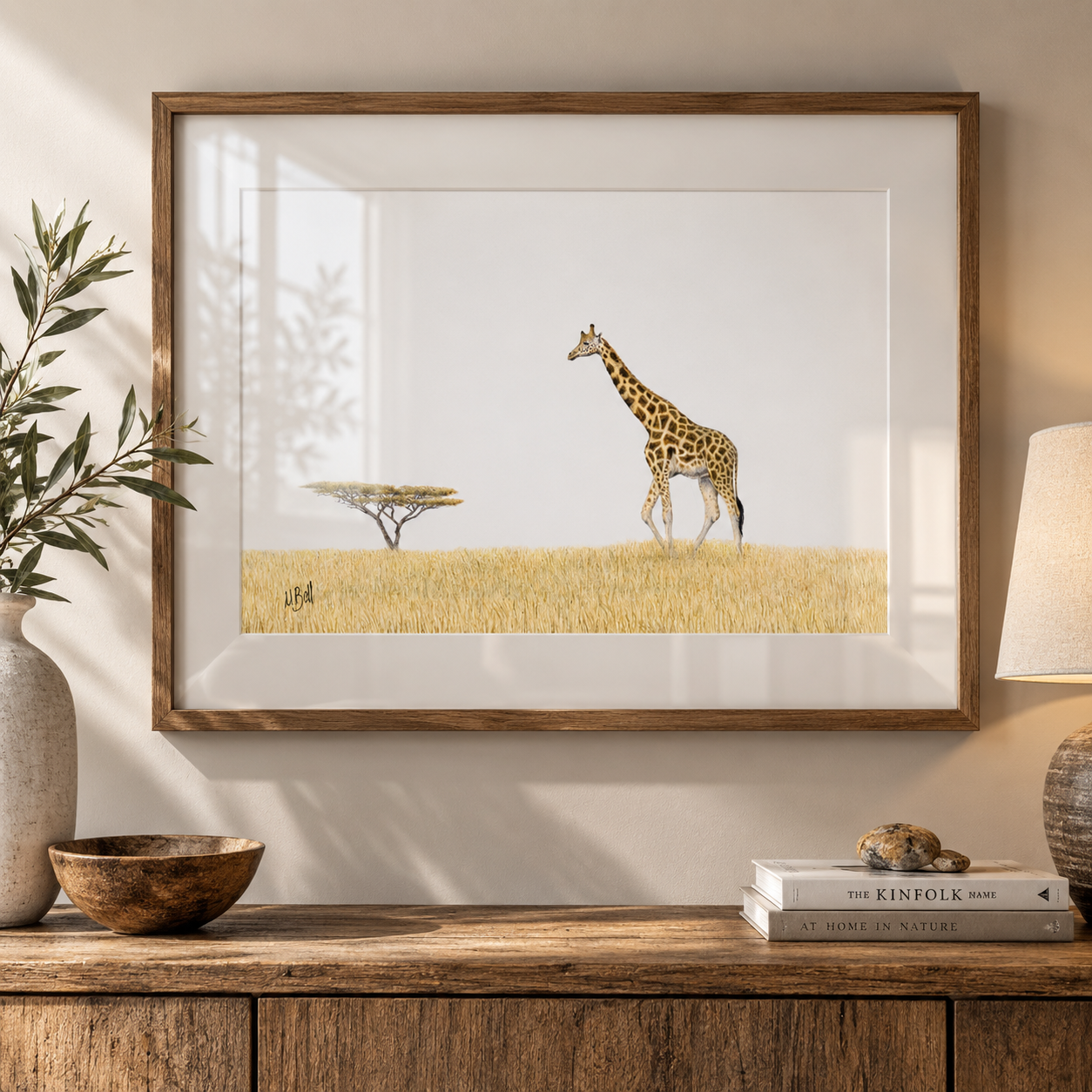 A framed art print depicting a giraffe and acacia tree on a golden savanna, styled on a wooden console table with a vase, bowl, and lamp.