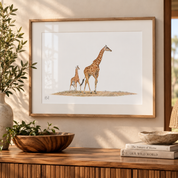 A framed art print featuring an illustration of a mother and child giraffe, styled on a wooden console table with a plant, bowls, and books in a warm, sunlit room.