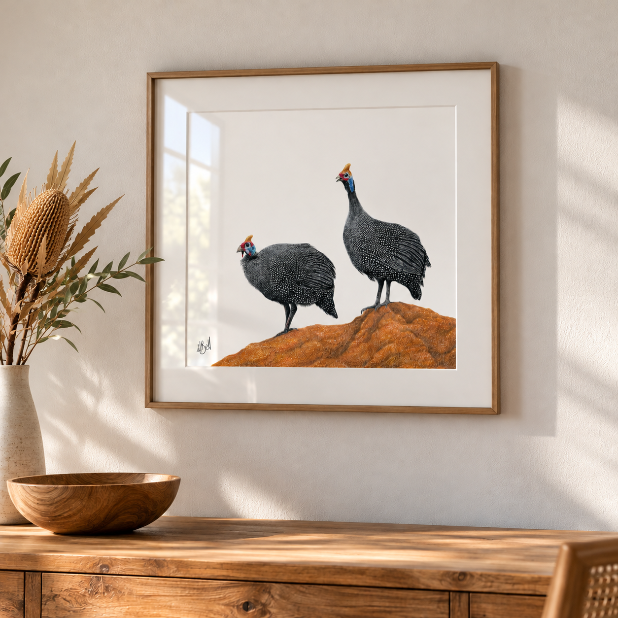 Framed artwork of two helmeted guineafowl on a rocky outcrop by South African wildlife artist Matthew Bell, displayed in a natural wood box frame with glass reflection in a warm, modern interior.