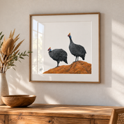 Framed artwork of two helmeted guineafowl on a rocky outcrop by South African wildlife artist Matthew Bell, displayed in a natural wood box frame with glass reflection in a warm, modern interior.