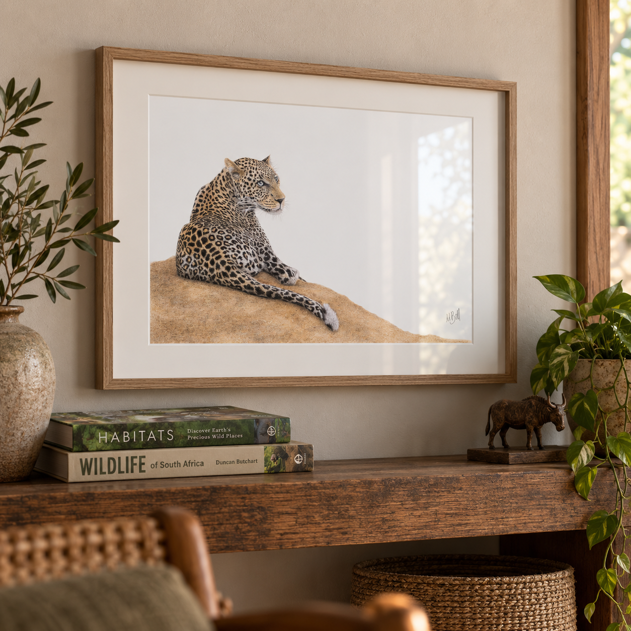 A framed art print of a leopard resting on a sandy hill, displayed on a wooden console table in a styled room setting with books, plants, and a small buffalo figurine.