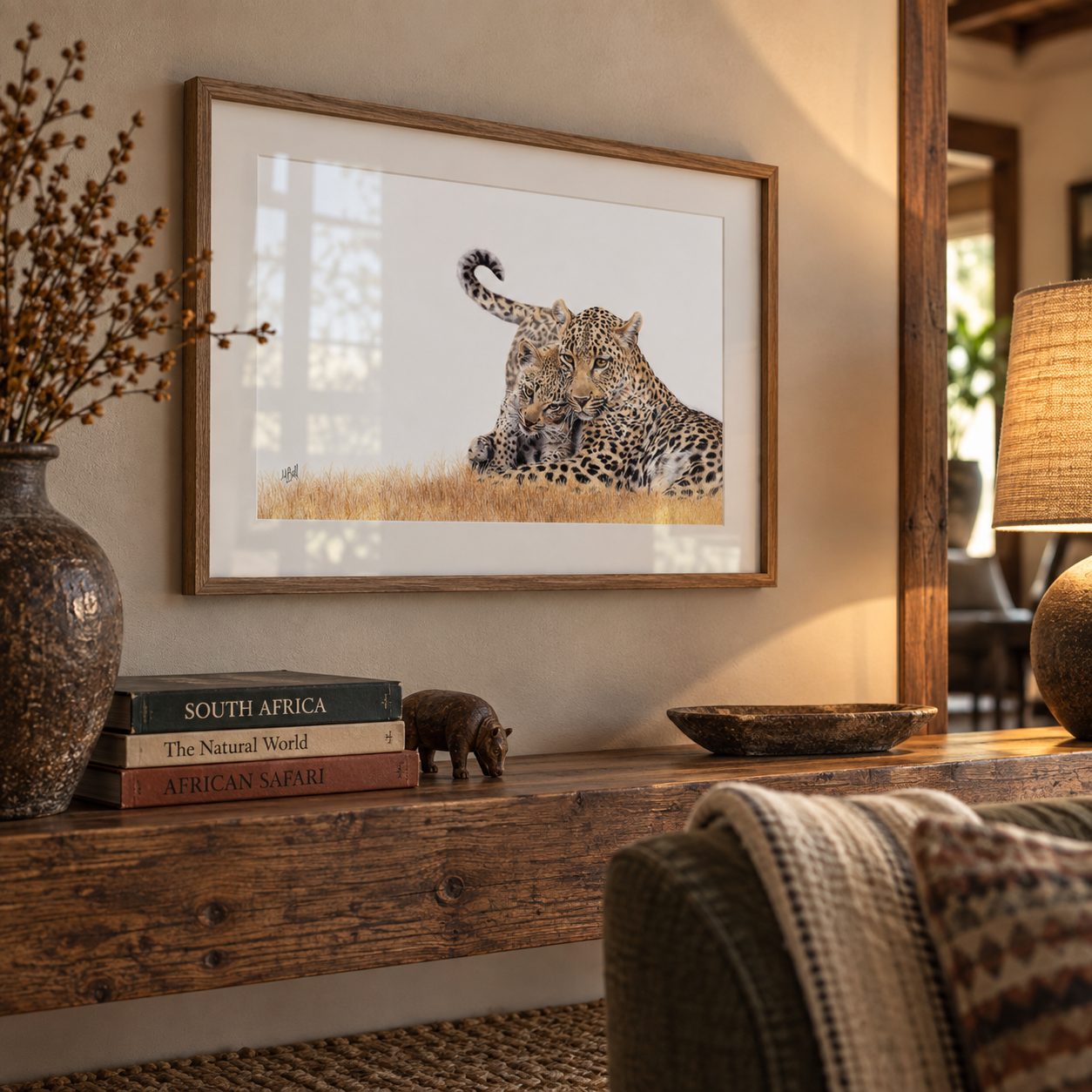 A framed art print of a leopard resting in a grassy landscape, displayed on a rustic wooden console table in a warm, styled room setting with books, a vase, and a lamp.