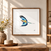 A framed art print of a Lilac Breasted Roller in flight, displayed on a warm beige wall above a rustic wooden table with a vase of yellow flowers.