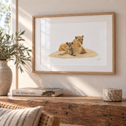 A framed art print depicting two lionesses resting on a sandy dune, displayed on a rustic wooden console table in a warm, sunlit room next to a potted plant and a book.