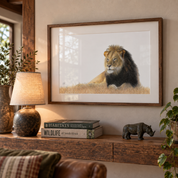 A framed art print of a lion resting in a grassy field, displayed on a wooden console table in a styled living room setting with a lamp, books, and a small rhinoceros figurine.