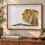 A framed lion artwork hangs on a beige wall above a wooden console table. The print shows a detailed lion with golden-brown fur on a white background, enclosed in a light wood frame.