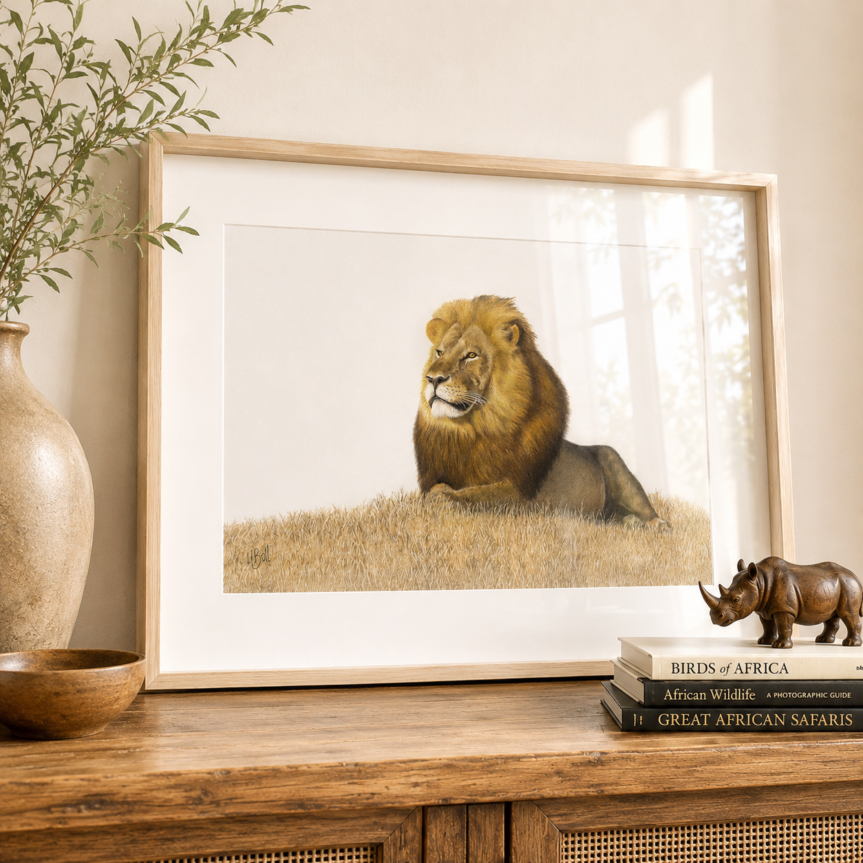 A framed art print of a male lion drawing by South African wildlife artist Matthew Bell, resting in a grassy landscape, displayed on a wooden console table next to a ceramic vase with greenery and a small rhinoceros figurine.