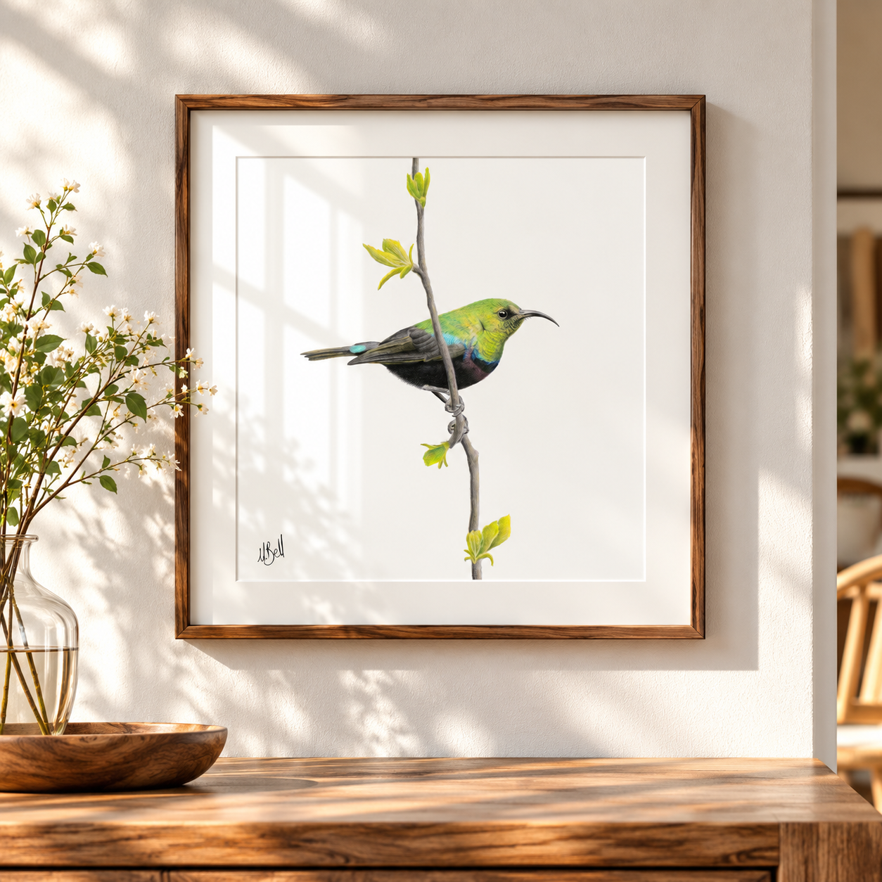 A framed Marico Sunbird print featuring a green and black bird perched on a branch, displayed on a wooden console table in a sunlit room with a vase of flowers nearby.