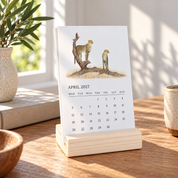 A vertical desk calendar standing on a light wood block stand on a wooden table, with a printed April 2027 page showing two illustrated cheetahs. The scene is styled in a sunlit room with a plant, books, and ceramic cups in the background.