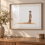 Meerkat artwork with an ash frame hanging on the wall with a vase of flowers next to it in a sunlit room